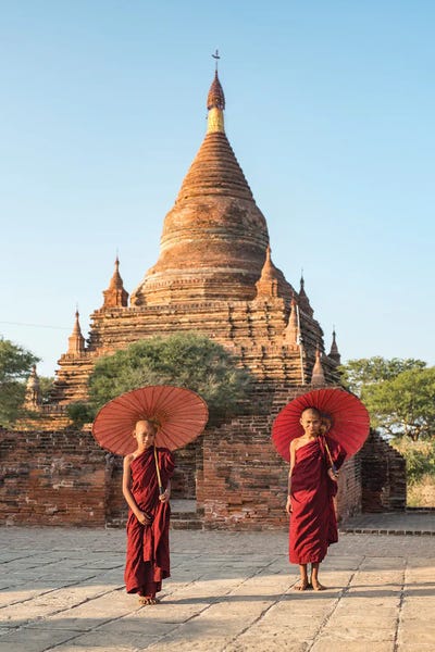 Buddhism: Two Young Novice Monks With Red Umbrellas Standing In Front Of A Temple, Bagan, Myanmar by Jan Becke