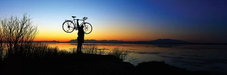 Silhouetted cyclist holding bicycle over head, river's edge, sunset, Alaska.