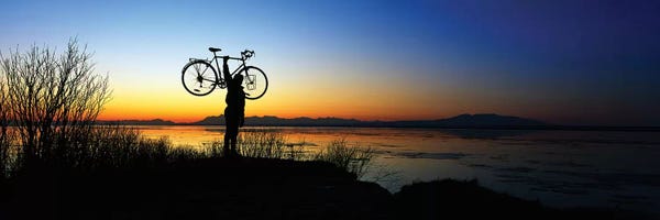 Alaska: Silhouetted cyclist holding bicycle over head, river's edge, sunset, Alaska. by Panoramic Images