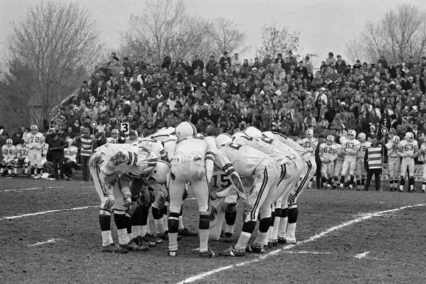 1980s Football Huddle On Playing Field With Spectators In Bleachers In The Background Outside