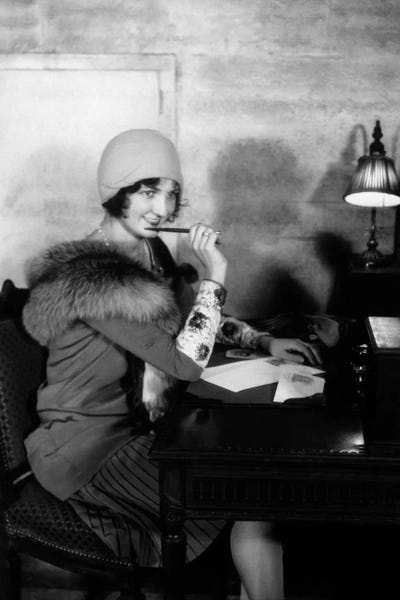 Historical Fashion: 1920s Smiling Woman With Pen To Lips Wearing Cloche Hat And Fur Collar Coat Writing A Letter At Hotel Lobby Desk With Lamp by Vintage Images