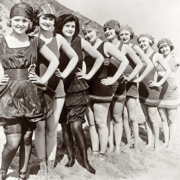 Historical Fashion: 1920s Group Of Smiling Women Wearing One Piece Bathing Suits And Caps Posing Lined Up On Beach Looking At Camera by Vintage Images