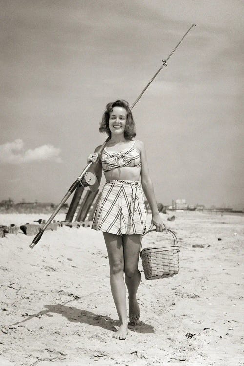 1940s Smiling Young Woman Walking On Beach Looking At Camera Wearing Two Piece Bathing Suit Skirt Carrying Surf Fishing Gear