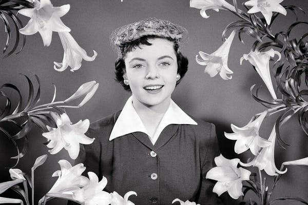 Historical Fashion: 1950s Portrait Smiling Brunette Woman Wearing Easter Bonnet Hat Looking At Camera Surrounded Framed By Easter Lilies by Vintage Images