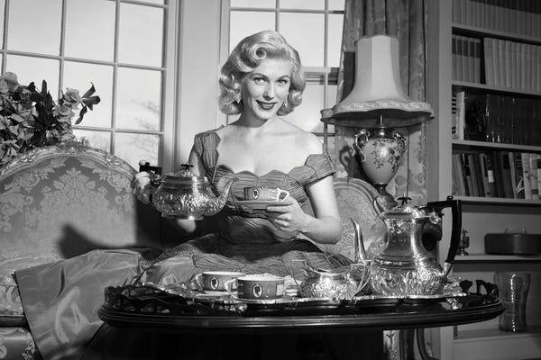 Historical Fashion: 1950s Smiling Blond Woman Elegant Dress & Home Furnishings Pouring Cup Tea From Silver Service Sitting Couch Looking At Camera by Vintage Images