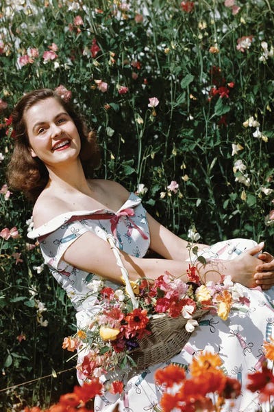 Historical Fashion: 1950s Smiling Woman Sitting In Meadow Holding Basket Of Flowers Looking At Camera by Vintage Images