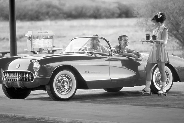 Historical Fashion: 1980s 1990s Carhop On Roller Skates Serving Drinks To Couple In Old Corvette Convertible At 1950s Style Drive-In Restaurant by Vintage Images